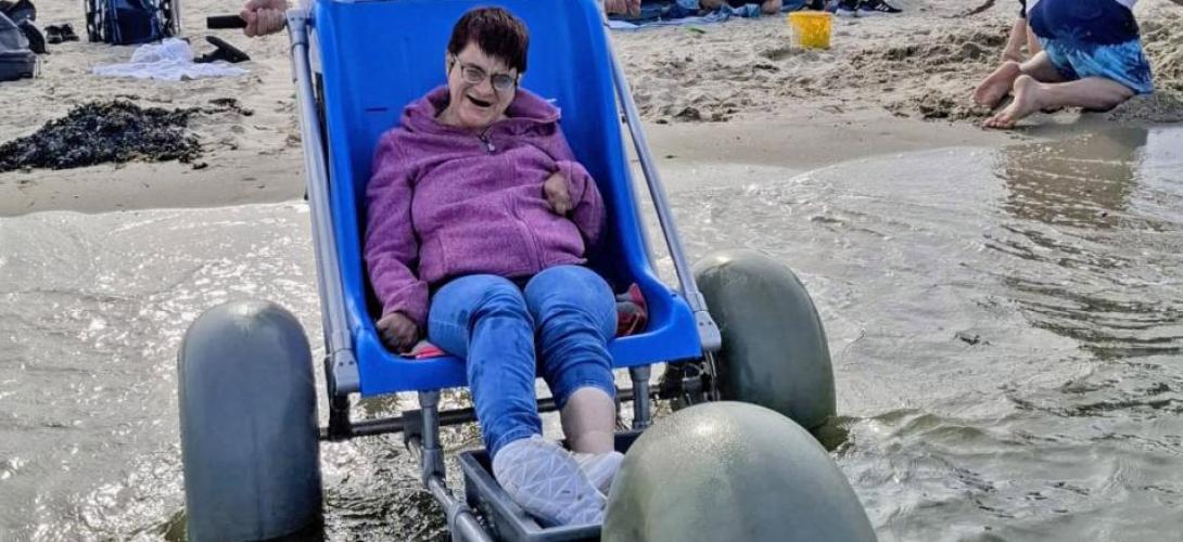 Ein Bild am Nordseestrand in Holland. Eine Frau sitzt am Meer an der Wasserkante im Strandrolli mit großen Ballonreifen. Sie lacht in die Kamera.