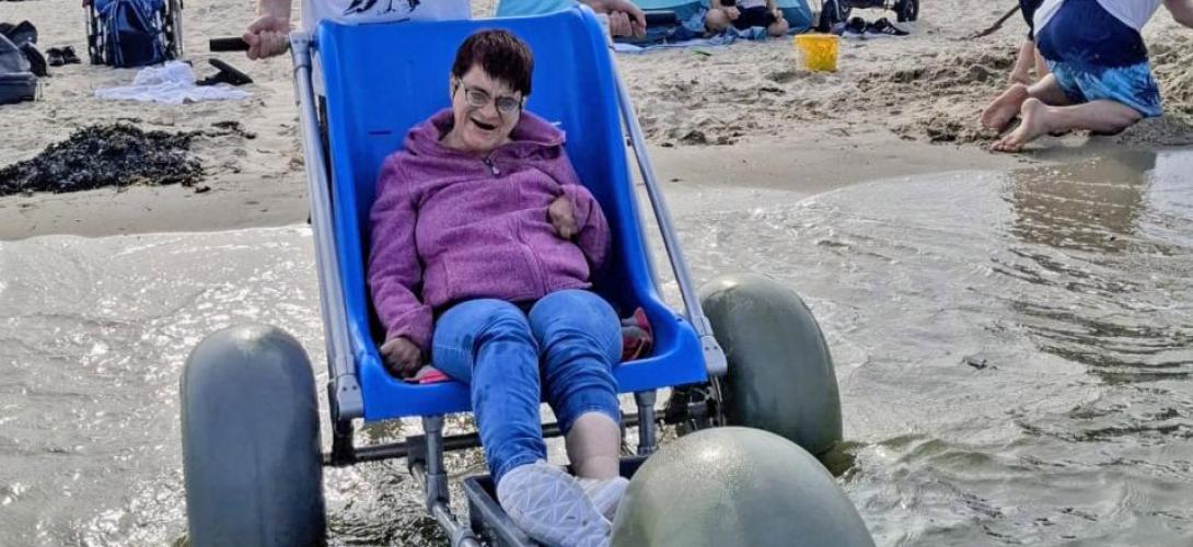 Ein Bild am Nordseestrand in Holland. Eine Frau sitzt am Meer an der Wasserkante im Strandrolli mit großen Ballonreifen. Sie lacht in die Kamera.