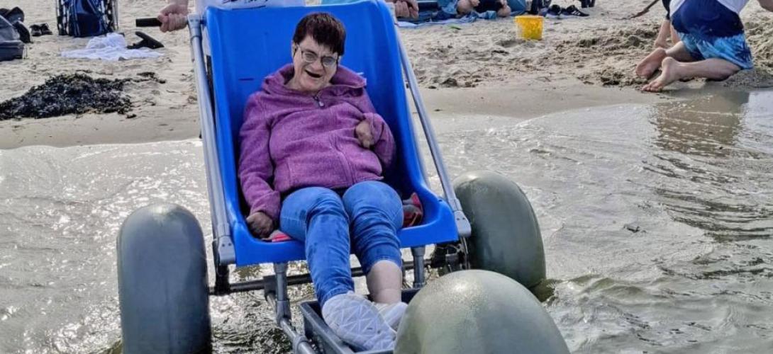 Ein Bild am Nordseestrand in Holland. Eine Frau sitzt am Meer an der Wasserkante im Strandrolli mit großen Ballonreifen. Sie lacht in die Kamera.