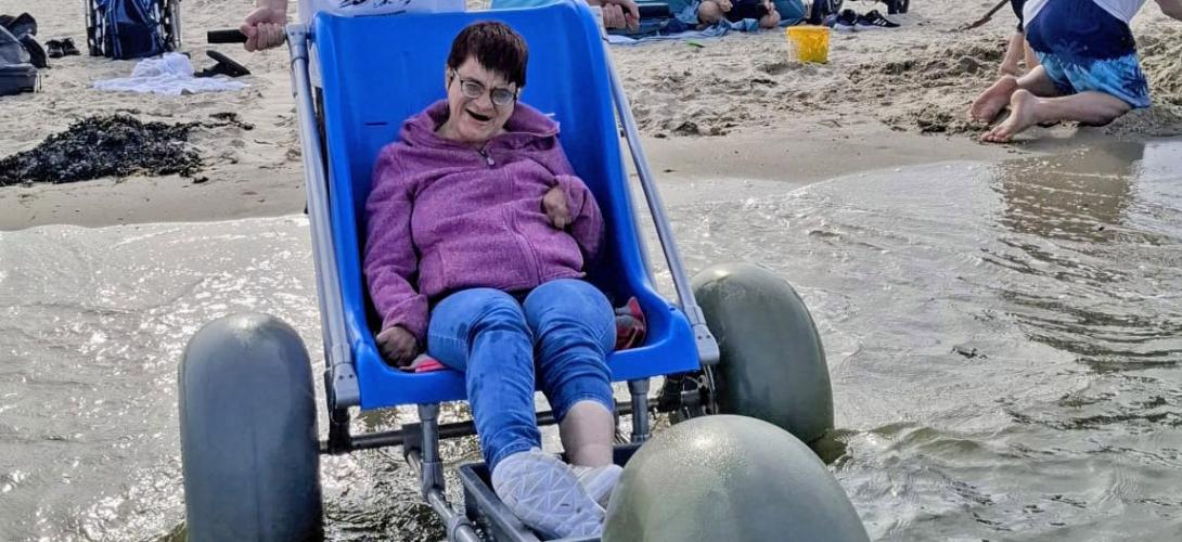 Ein Bild am Nordseestrand in Holland. Eine Frau sitzt am Meer an der Wasserkante im Strandrolli mit großen Ballonreifen. Sie lacht in die Kamera.