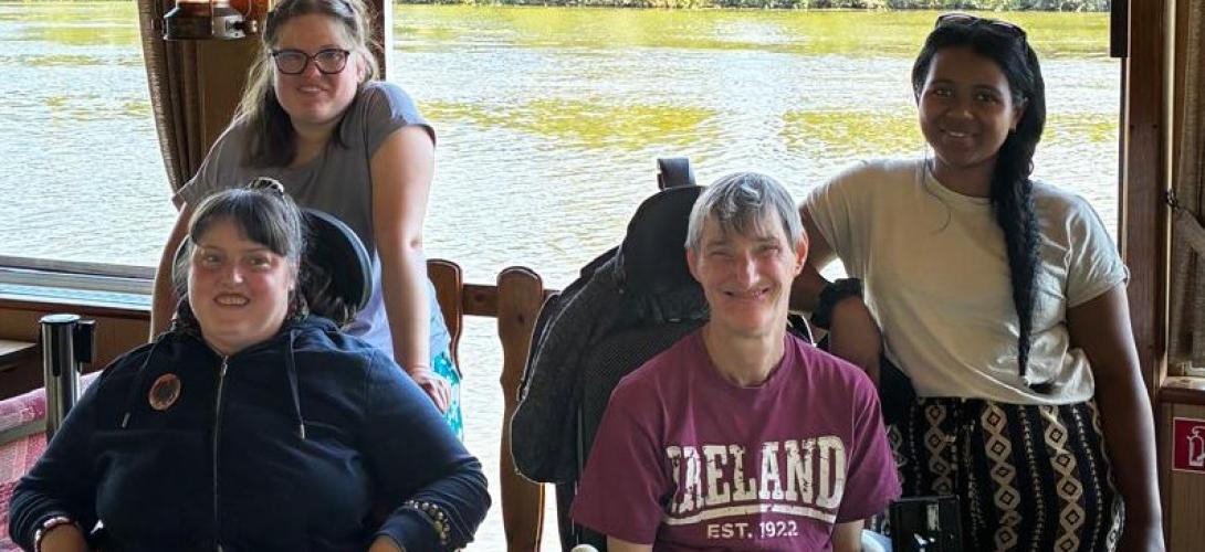 Zu sehen sind vier Frauen, zwei davon im Rollstuhl. Sie lachen in die Kamera. Im Hintergrund fließt ein Fluss.