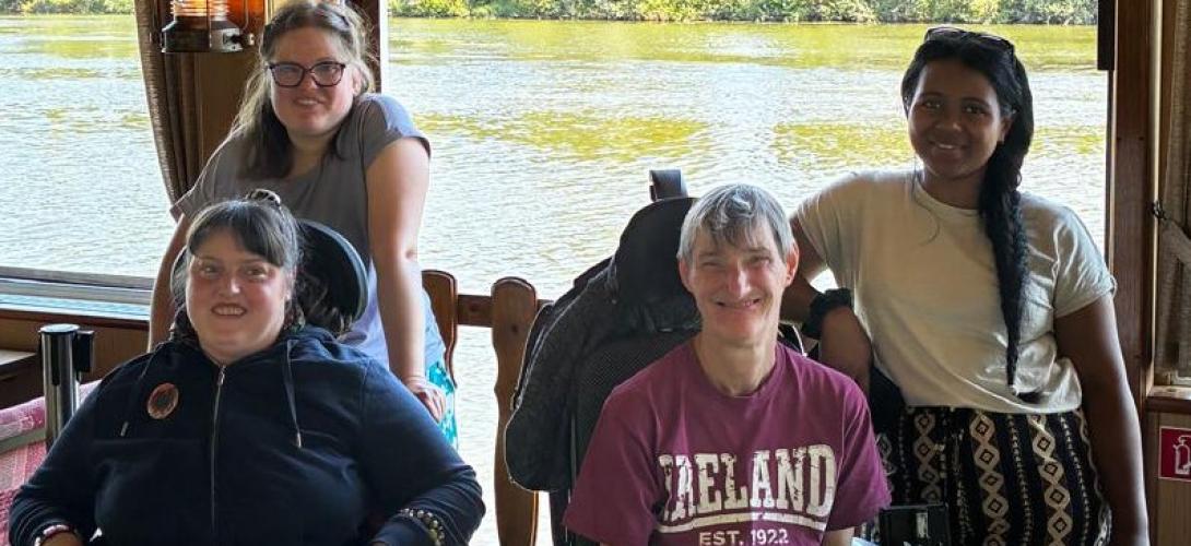 Zu sehen sind vier Frauen, zwei davon im Rollstuhl. Sie lachen in die Kamera. Im Hintergrund fließt ein Fluss.