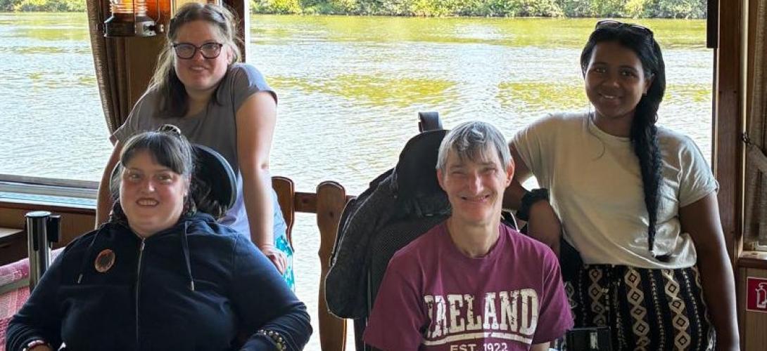 Zu sehen sind vier Frauen, zwei davon im Rollstuhl. Sie lachen in die Kamera. Im Hintergrund fließt ein Fluss.